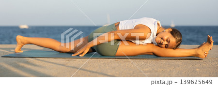 On windy day young slender woman does yoga near sea doing Hanumasana 139625649