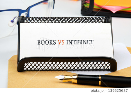 White card showing BOOKS VS INTERNET in a black desk holder. A sleek fountain pen lies on a brown surface. Blue eyeglasses and colorful sticky notes are in the background White card showing BOOKS VS INTERNET in a black desk holder. A sleek fountain pen lies on a brown surface. Blue eyeglasses and colorful sticky notes are in the background 139625687