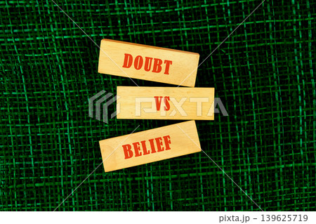 Three wooden blocks vertically stack displaying the words Doubt, VS, and Belief in red capital letters on a dark green textured surface. This illustrates a conceptual conflict 139625719