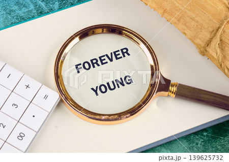 A magnifying glass highlights the phrase Forever Young written on a clean white page. A white calculator and a piece of natural wood are also on the desk 139625732
