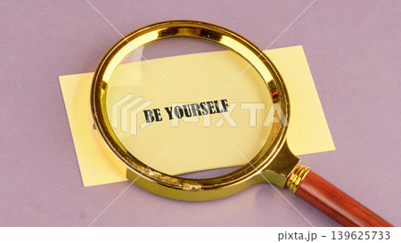 A golden magnifying glass with a brown handle rests over a yellow note, clearly showing the bold words be yourself 139625733
