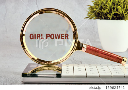 A magnifying glass with a wooden handle rests on a white keyboard. It sharply focuses on the phrase Girl Power. A green potted plant stands in the background on a grey desk A magnifying glass with a wooden handle rests on a white keyboard. It sharply focuses on the phrase Girl Power. A green potted plant stands in the background on a grey desk 139625741
