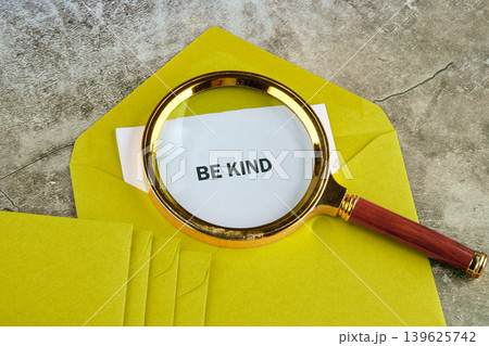 A magnifying glass rests on a white note card inside an open bright yellow envelope. The note displays the clear words BE KIND A magnifying glass rests on a white note card inside an open bright yellow envelope. The note displays the clear words BE KIND 139625742