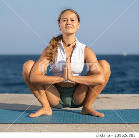 On windy day young slender woman does yoga near sea doing Malasana On windy day young slender woman does yoga near sea doing Malasana 139626885