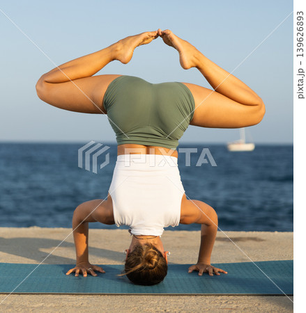 Sportive girl practicing headstand pose of yoga on seashore Sportive girl practicing headstand pose of yoga on seashore 139626893