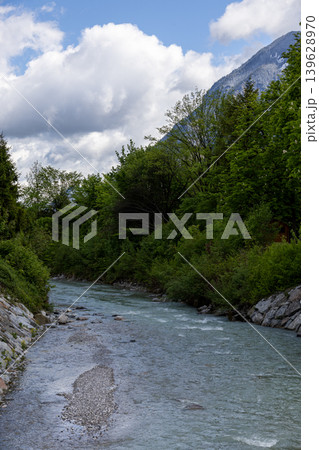Alpine River Flowing Through Lush Green Forest Beneath a Cloudy Spring Sky Alpine River Flowing Through Lush Green Forest Beneath a Cloudy Spring Sky 139628970