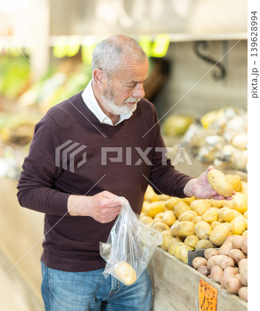 Elderly man visiting a vegetable store stands near the counter and chooses farm potato Elderly man visiting a vegetable store stands near the counter and chooses farm potato 139628994