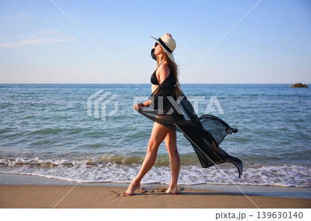 Woman walking along sandy beach in black bikini with flowing cover up 139630140