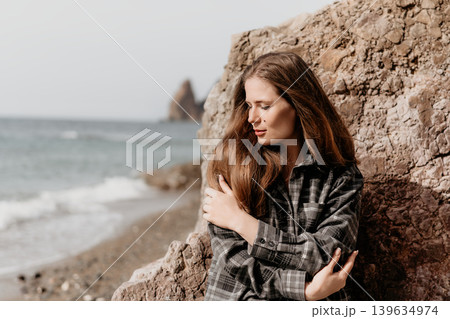 Woman beach portrait pensive female hugging herself by rock formation at the shore in contemplation during peaceful day Woman beach portrait pensive female hugging herself by rock formation at the shore in contemplation during peaceful day 139634974
