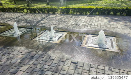 A park fountain with jets of water, surrounded by green trees. 139635477