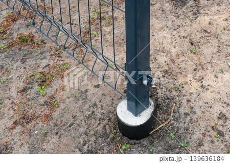 Metal fence post fixed in concrete foundation with wire mesh panel on sandy ground closeup image 139636184