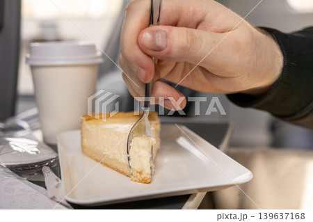 Slice of creamy cheesecake served on a tray during a flight with a person using a silver spoon to eat the dessert, representing comfort, travel dining, and leisure during air transport trips 139637168