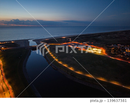 Dramatic aerial night view of a Dutch coastal landscape featuring a calm waterway canal flowing toward the North Sea. Warm streetlights illuminate roads and a parking area at twilight. Dramatic aerial night view of a Dutch coastal landscape featuring a calm waterway canal flowing toward the North Sea. Warm streetlights illuminate roads and a parking area at twilight. 139637381