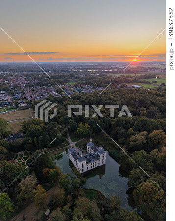 Majestic aerial sunset over a historic moated castle surrounded by dense forest. Golden hour light illuminates the castle architecture and the sprawling European town and countryside beyond. 139637382