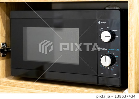 Black microwave oven installed in a wooden kitchen cabinet, featuring manual dial controls for power and timer settings in a modern domestic cooking and food preparation environment Black microwave oven installed in a wooden kitchen cabinet, featuring manual dial controls for power and timer settings in a modern domestic cooking and food preparation environment 139637434