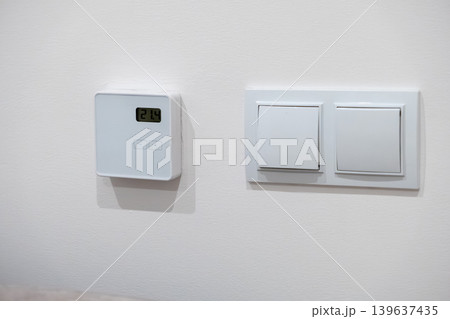 Digital wall thermostat with light switch on white surface, representing modern climate control technology and energy efficient home automation for comfortable living environments Digital wall thermostat with light switch on white surface, representing modern climate control technology and energy efficient home automation for comfortable living environments 139637435