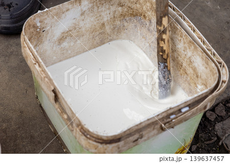 Old plastic bucket containing white wall paint being stirred with a wooden stick to ensure smooth texture for renovation or home improvement projects in a residential setting 139637457