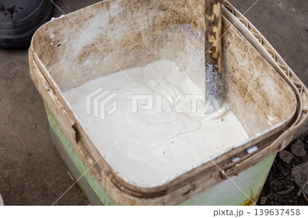 White construction paint being mixed in a weathered plastic bucket with a wooden stick, symbolizing professional renovation, home improvement, DIY repair work, and maintenance tasks White construction paint being mixed in a weathered plastic bucket with a wooden stick, symbolizing professional renovation, home improvement, DIY repair work, and maintenance tasks 139637458