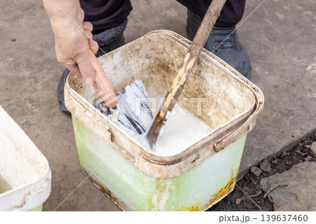 Person mixing white lime mortar in a plastic bucket using a metal trowel and wooden stick, representing preparation of materials for masonry work, plastering, or renovation task 139637460