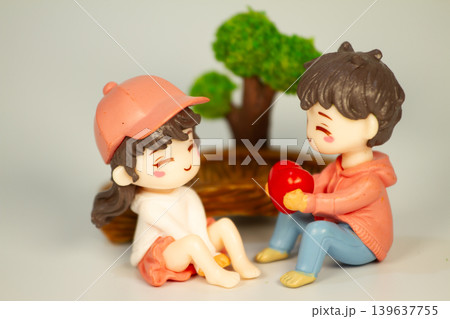 Adorable miniature couple figurines sharing a sweet heart gift moment beside a decorative tree and wooden bridge in a playful romantic setting. Adorable miniature couple figurines sharing a sweet heart gift moment beside a decorative tree and wooden bridge in a playful romantic setting. 139637755