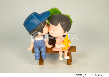 Sweet miniature boy and girl figurines sharing a kiss on a wooden bench while holding a yellow teddy bear in a romantic decorative scene. Sweet miniature boy and girl figurines sharing a kiss on a wooden bench while holding a yellow teddy bear in a romantic decorative scene. 139637756