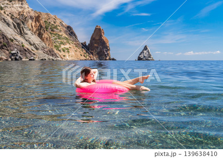 Woman floating inflatable ring in crystal clear sea water near rocky cliffs during sunny summer vacation Woman floating inflatable ring in crystal clear sea water near rocky cliffs during sunny summer vacation 139638410