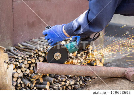 A worker in blue gloves uses an electric angle grinder to cut a metal pipe, creating bright sparks during a professional repair project to demonstrate industrial steel labor today A worker in blue gloves uses an electric angle grinder to cut a metal pipe, creating bright sparks during a professional repair project to demonstrate industrial steel labor today 139638692