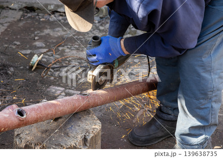 An industrial worker carefully cuts through a thick rusty steel pipe with an angle grinder while bright orange sparks fly across the dark workshop during a professional metal fabrication job An industrial worker carefully cuts through a thick rusty steel pipe with an angle grinder while bright orange sparks fly across the dark workshop during a professional metal fabrication job 139638735
