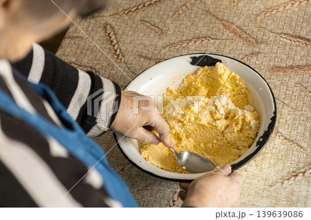 An elderly woman stirs a yellow cottage cheese mixture in an enamel bowl, preserving traditional home cooking methods and family heritage within an authentic, rustic kitchen culinary space An elderly woman stirs a yellow cottage cheese mixture in an enamel bowl, preserving traditional home cooking methods and family heritage within an authentic, rustic kitchen culinary space 139639086