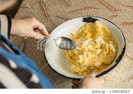 An elderly person stirs fresh cottage cheese and a raw egg in a metal bowl while preparing traditional dairy ingredients for authentic healthy family baking in a cozy rustic home kitchen An elderly person stirs fresh cottage cheese and a raw egg in a metal bowl while preparing traditional dairy ingredients for authentic healthy family baking in a cozy rustic home kitchen 139639087