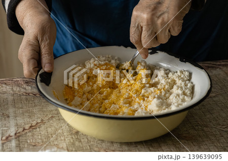 An elderly person carefully mixes cottage cheese and egg yolk in a bowl to prepare dough for traditional homemade pastries, reflecting the patience and art of classic rustic home cooking An elderly person carefully mixes cottage cheese and egg yolk in a bowl to prepare dough for traditional homemade pastries, reflecting the patience and art of classic rustic home cooking 139639095