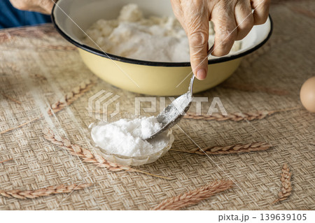 An elderly woman adds sugar with a metal spoon into a bowl of cottage cheese for a homemade recipe, highlighting traditional baking processes, rustic kitchen aesthetics, and healthy meals An elderly woman adds sugar with a metal spoon into a bowl of cottage cheese for a homemade recipe, highlighting traditional baking processes, rustic kitchen aesthetics, and healthy meals 139639105