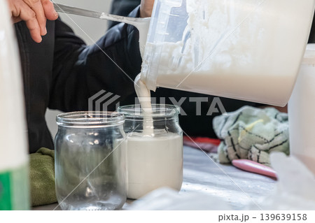 A professional chef carefully pours fresh, artisanal sour cream from a container into a glass jar for retail packaging or culinary preparation in a clean, high-quality commercial kitchen 139639158