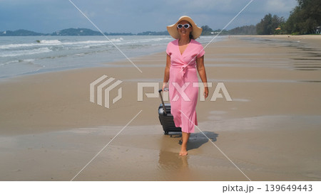 Young barefoot woman in sunglasses, pink dress and straw hat pulling a suitcase along sandy beach enjoying sea view. Vacation leisure concept Young barefoot woman in sunglasses, pink dress and straw hat pulling a suitcase along sandy beach enjoying sea view. Vacation leisure concept 139649443