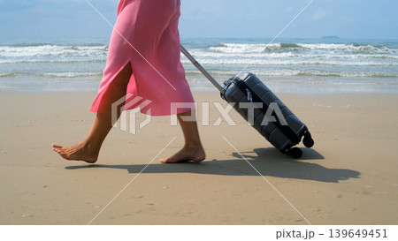 Young barefoot woman in pink dress rolling a grey suitcase along sandy beach with blue sea and waves in background. Summer travel concept Young barefoot woman in pink dress rolling a grey suitcase along sandy beach with blue sea and waves in background. Summer travel concept 139649451