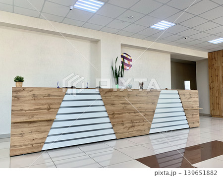 Modern reception desk showcases a wooden finish with geometric patterns in a bright office space, complemented by indoor plants and decorative items 139651882