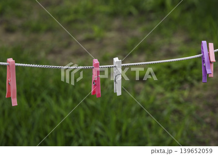 A row of pink, white, and purple plastic pegs attached to a white rope. The background shows a blurred garden with green grass and soil during a bright day. A row of pink, white, and purple plastic pegs attached to a white rope. The background shows a blurred garden with green grass and soil during a bright day. 139652059