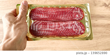 Hand holding a pack of sliced salami on a wooden table mid day Hand holding a pack of sliced salami on a wooden table mid day 139652890