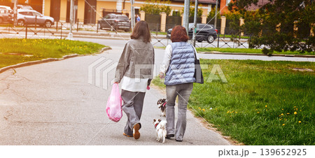 Two women walk together along a path with a small dog in a city at dusk 139652925