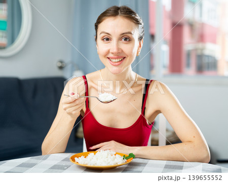 Young woman eating rice while sitting at the table in room. Diet and weight loss concept Young woman eating rice while sitting at the table in room. Diet and weight loss concept 139655552
