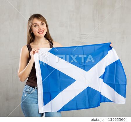 Happy young woman holding flag of Scotland against unicoloured background Happy young woman holding flag of Scotland against unicoloured background 139655604