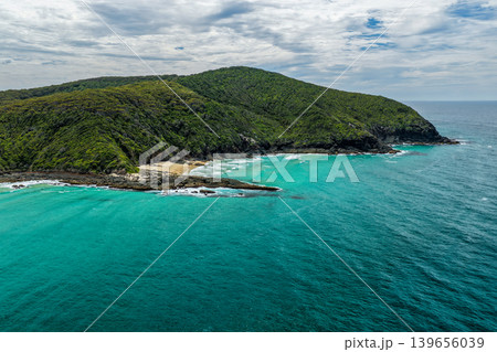 Aerial drone view of Sharkies Beach featuring a rugged rocky coastline and a tree covered mountain backdrop. 139656039