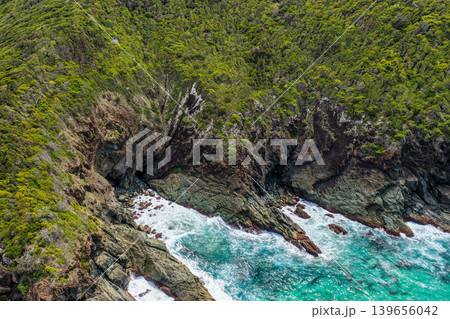 Aerial drone view of Sharkies Beach featuring a rugged rocky coastline and a tree covered mountain backdrop. 139656042
