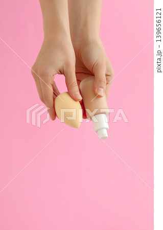 Hands hold a beauty sponge and a tube of cream against pink wall 139656121