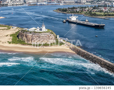 Aerial drone view of Nobbys Lighthouse and Nobbys Beach with a large cargo ship being escorted by tug boats through Newcastle Harbour in the background. 139656145