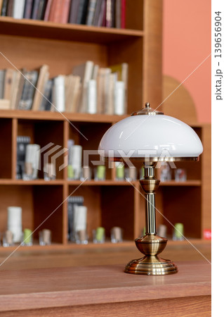 A stylish table lamp sits on a wooden surface beside a bookshelf filled with neatly arranged books, creating a cozy reading atmosphere in natural light. 139656904