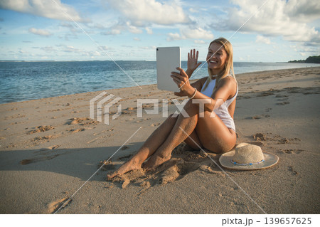 Woman making a video call while sitting on the sand Woman making a video call while sitting on the sand 139657625
