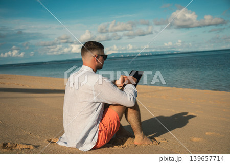 Person sitting on sand using a tablet by the sea Person sitting on sand using a tablet by the sea 139657714