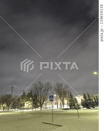A peaceful winter night scene shows a pedestrian crossing sign beneath bare trees, lit by streetlights under a cloudy sky. Perfect for themes of urban landscapes and winter safety. 139659138