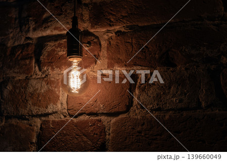A vintage filament light bulb emits a warm amber glow in front of brick wall A vintage filament light bulb emits a warm amber glow in front of brick wall 139660049